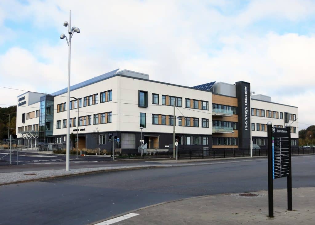 Rexuma vårdrumspanel Ett modernt trevåningssjukhus med stora fönster och vit fasad står vid en tom gata. En svart skylt med vit text står på framsidan, bredvid en hög gatlykta. Den delvis molniga himlen ramar in den eleganta Vårdrumspanelens design.