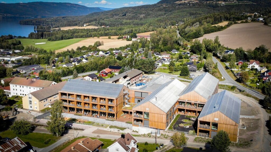Rexuma vårdrumspanel Flygfoto över moderna träbyggnader, bl.a. ett sjukhus med Vårdrumspanel, omgivna av hus, fält, träd och en sjö i ett kuperat jordbrukslandskap under klar himmel.