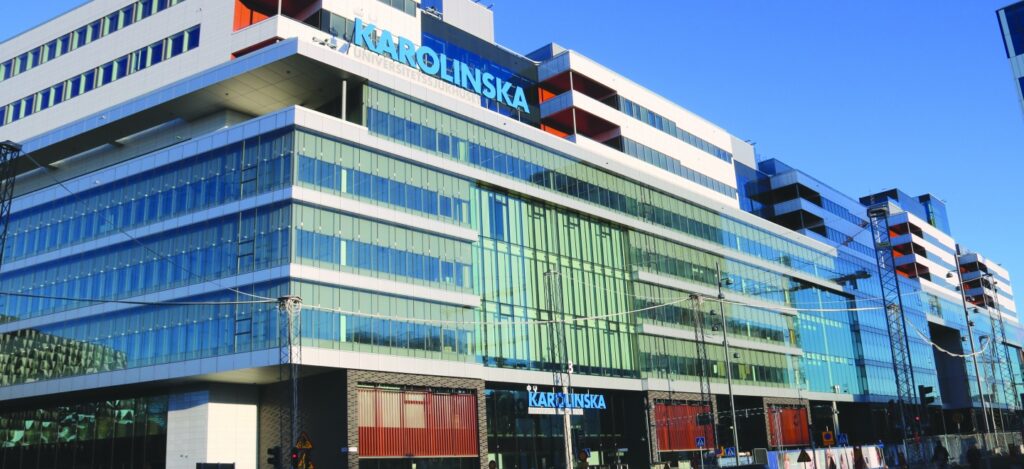 Rexuma vårdrumspanel En modern sjukhusbyggnad i glas och stål med ordet Karolinska på fasaden, som speglar en klarblå himmel. Flera våningar och stora fönster är synliga, med gatukablar i förgrunden.