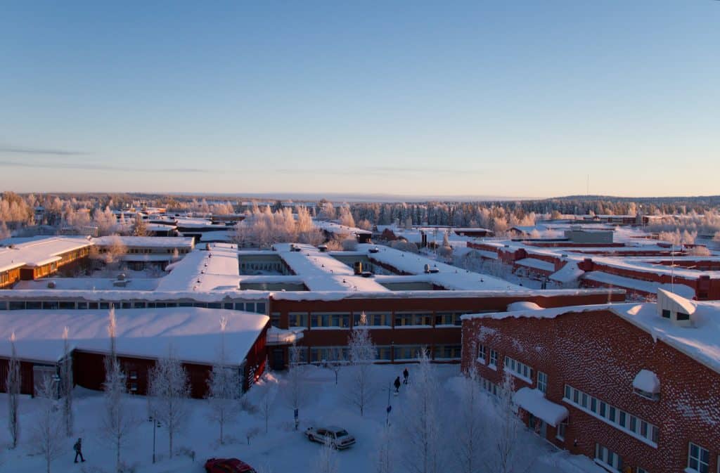 Rexuma vårdrumspanel Flygfoto över en snötäckt stad med röda tegelbyggnader, frostnupna träd och bilar parkerade längs snöiga gator. Ett sjukhus i närheten sticker ut under den klarblå himlen vid soluppgång eller solnedgång.