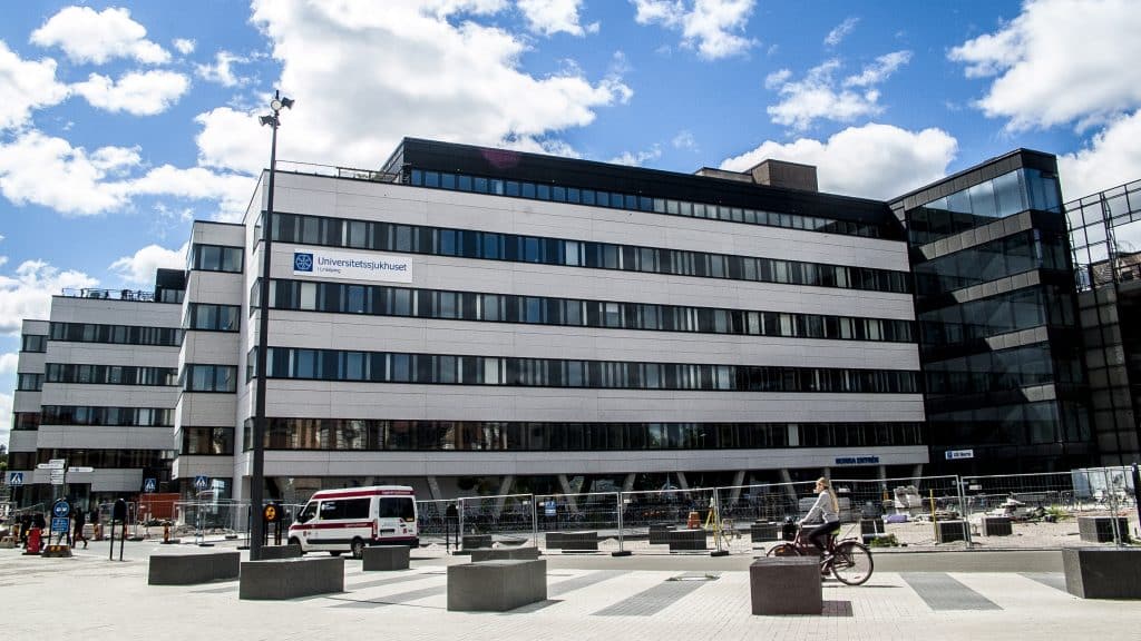 Rexuma vårdrumspanel En modern sjukhusbyggnad i flera våningar med vit och svart fasad med texten Universitetssjukhuset. En person cyklar och en vit skåpbil står parkerad utanför, under en delvis molnig blå himmel.