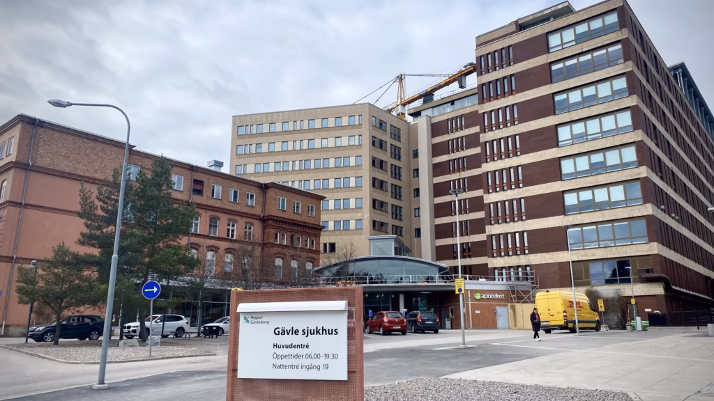 Rexuma vårdrumspanel Ett sjukhuskomplex med flera moderna byggnader, en svängd entré och en skylt med texten ”Gävle sjukhus”. Flera bilar och en gul skåpbil står parkerade utanför under en molnig himmel.