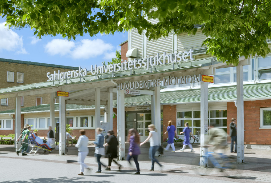 Rexuma vårdrumspanel Människor går förbi ingången till Sahlgrenska universitetssjukhuset i Sverige. Över ingången sitter en skylt, och i förgrunden syns träd med gröna blad. Scenen är ljus och livlig.