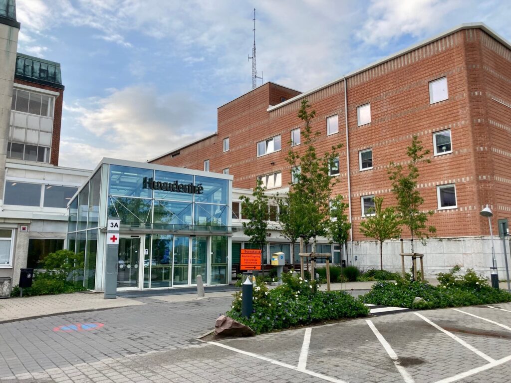 Rexuma vårdrumspanel En modern sjukhusentré med skylten ”Haukeland”, glasdörrar, en tegelbyggnad, träd, parkeringsplatser och sjukvårdsskyltar syns utifrån under en delvis molnig himmel.